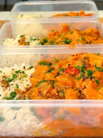 Three meal prep containers filled with creamy Tuscan tofu and rice, garnished with fresh herbs. The tofu is coated in a rich, orange-colored sauce, creating a visually appealing and appetizing dish.