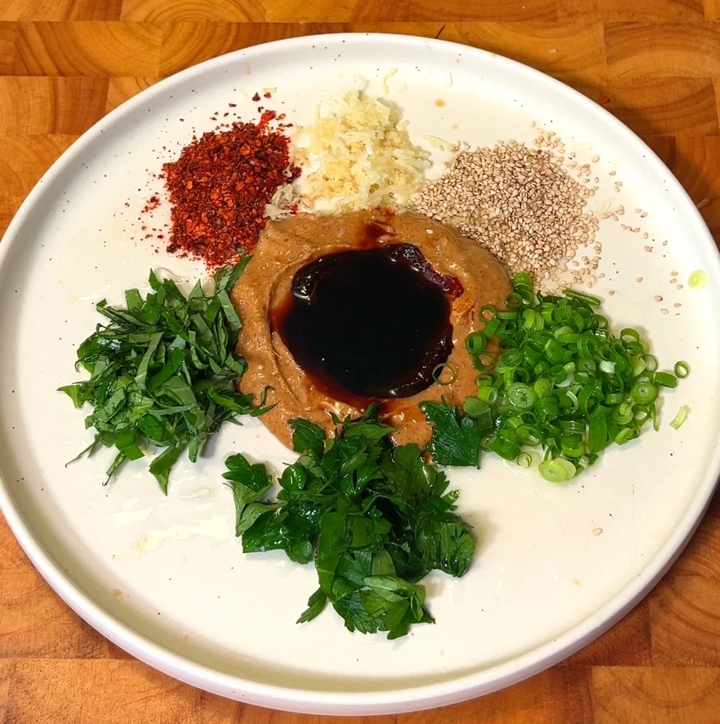 Whisking together a rich, spicy noodle sauce with chili flakes, garlic, and fresh herbs on a plate.