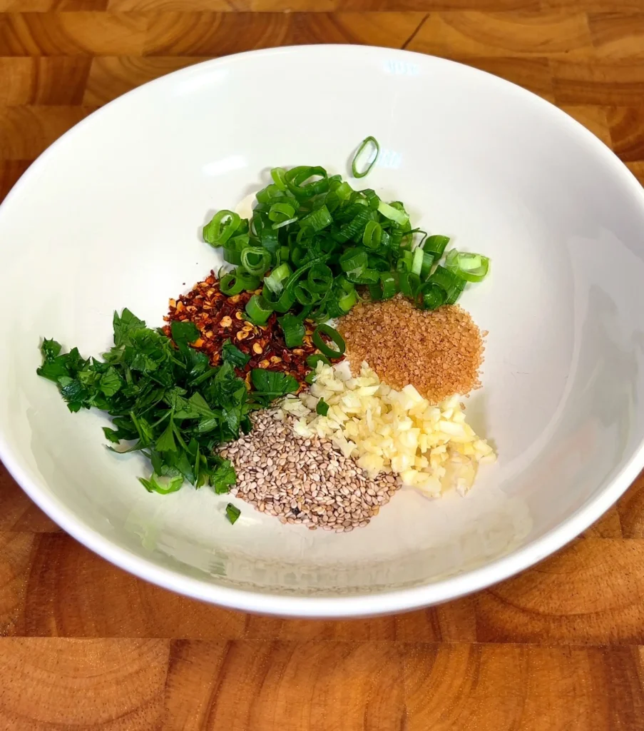 Fresh ingredients in a bowl for making spicy noodle sauce, including green onions, garlic, sesame seeds, chili flakes, and parsley