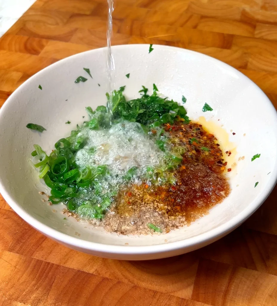 Hot water being poured into a bowl of spicy noodle sauce ingredients including green onions, sesame seeds, and chili oil