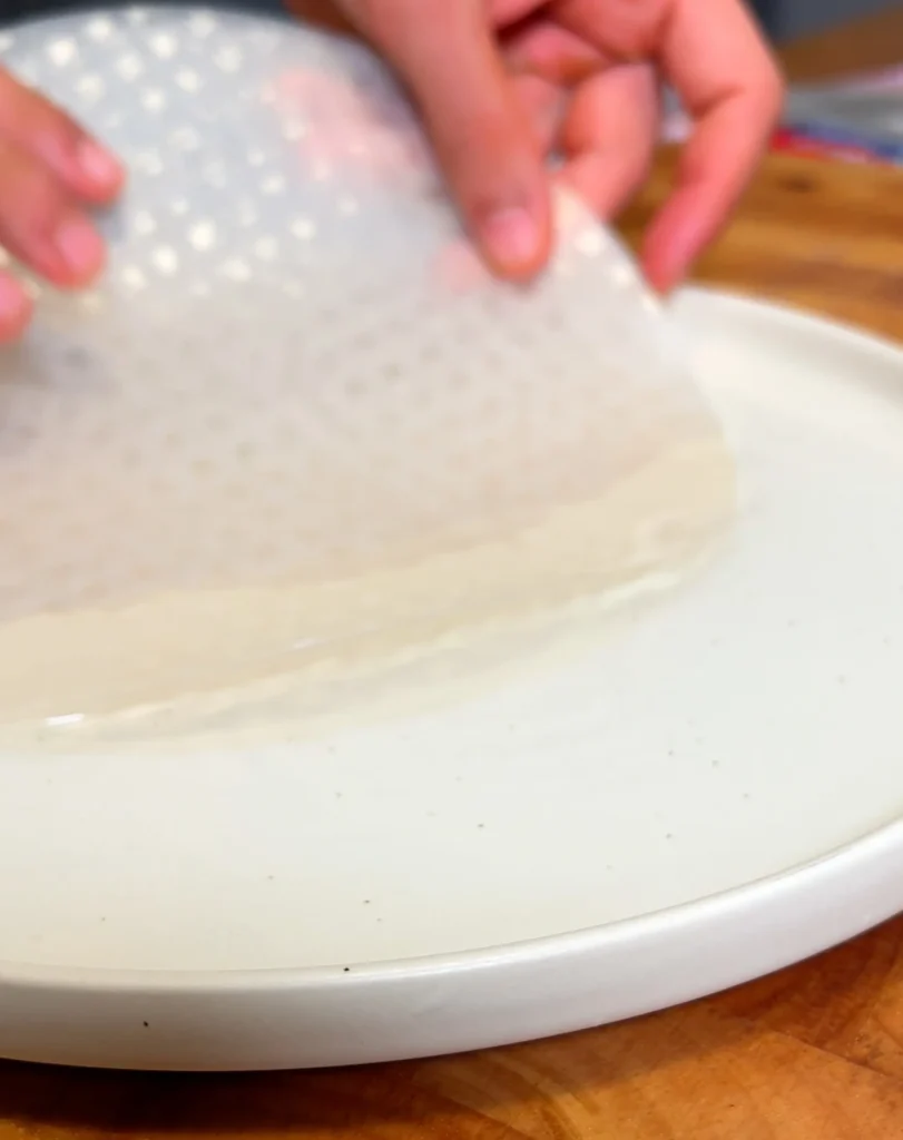 Hands preparing rice paper on a white plate for making fresh spring rolls.