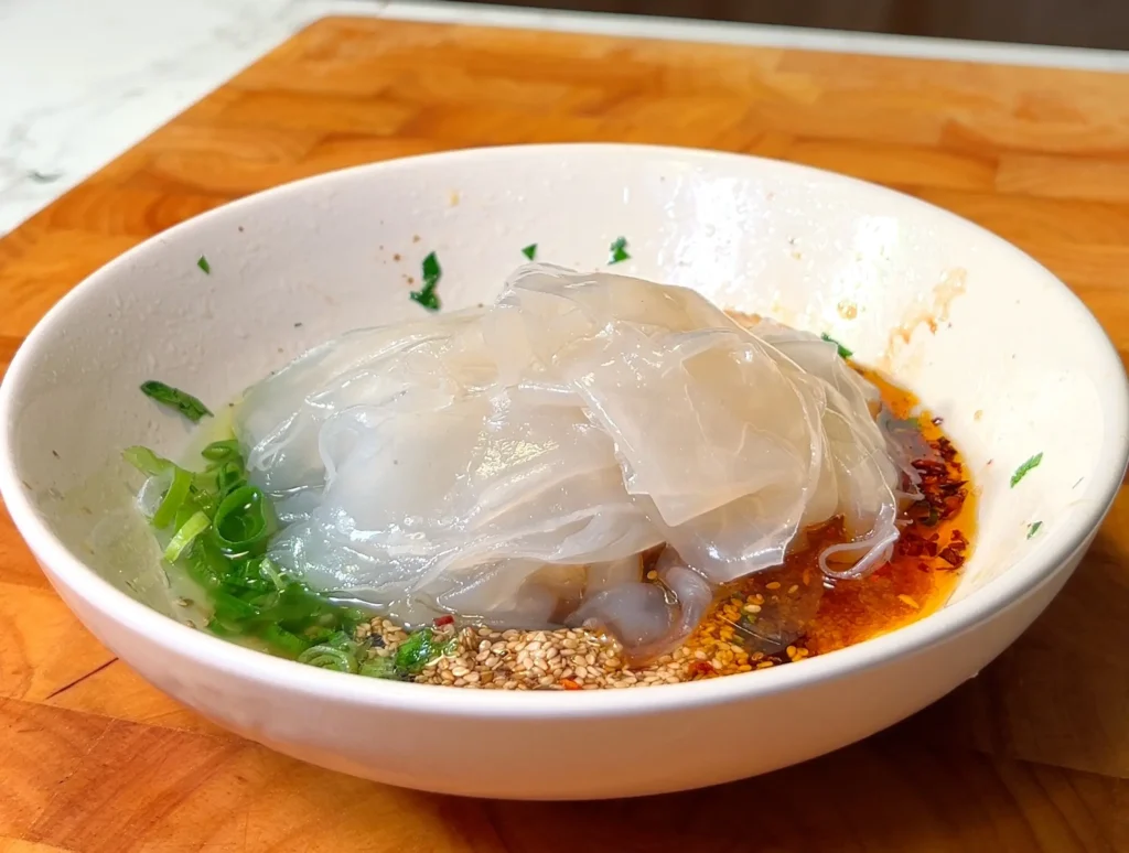 White bowl with freshly prepared rice noodles in a savory sauce topped with green onions, sesame seeds, and chili oil.