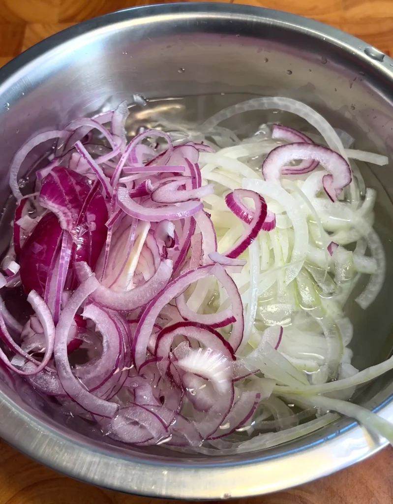 Thinly sliced red and white onions soaking in water in a metal bowl