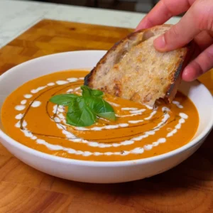 Creamy tomato soup garnished with fresh basil and drizzled with cream, served with a slice of crusty bread.