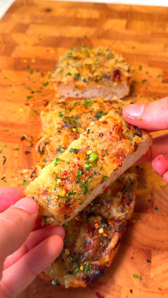 A close-up of a hand holding a slice of crispy, cheesy garlic bread garnished with fresh herbs and chili flakes, with more garlic bread slices in the background on a wooden cutting board.