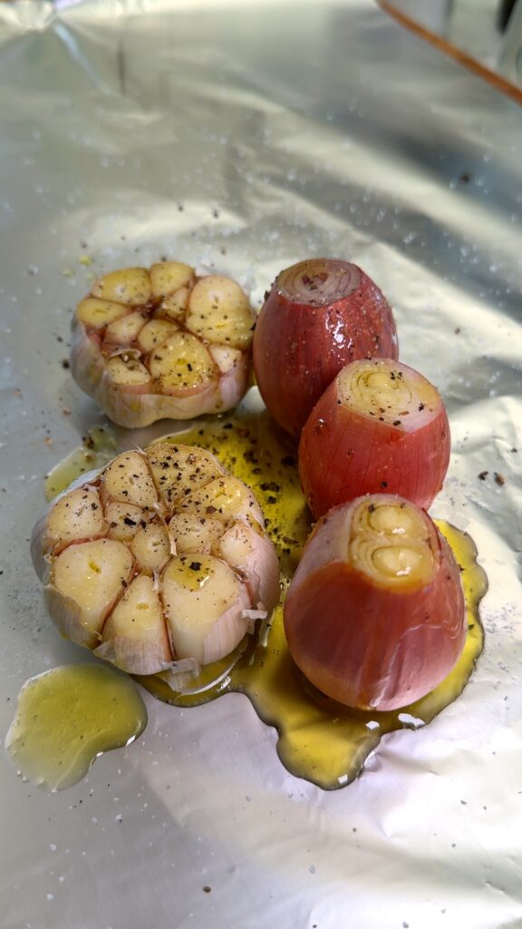 Halved garlic bulbs and shallots drizzled with olive oil, seasoned with salt and pepper, placed on aluminum foil, ready for roasting.