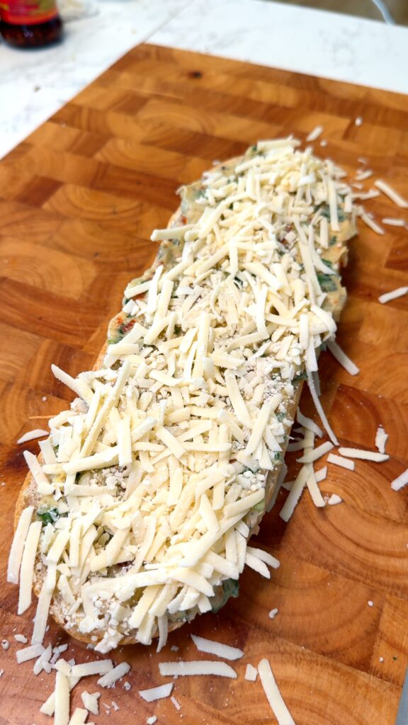 Unbaked garlic bread topped with a generous layer of shredded cheese and herb butter mixture, resting on a wooden cutting board.