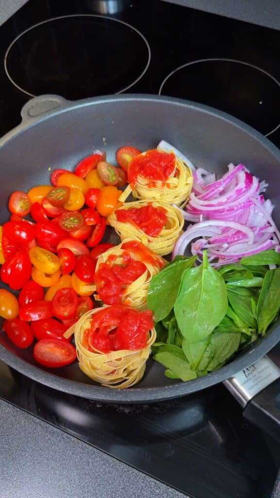Layer ingredients – In a large pan, place the pasta nests in the center. Arrange cherry tomatoes, onion, garlic, and spinach around. Add tomato puree and paste on top, then drizzle with olive oil.