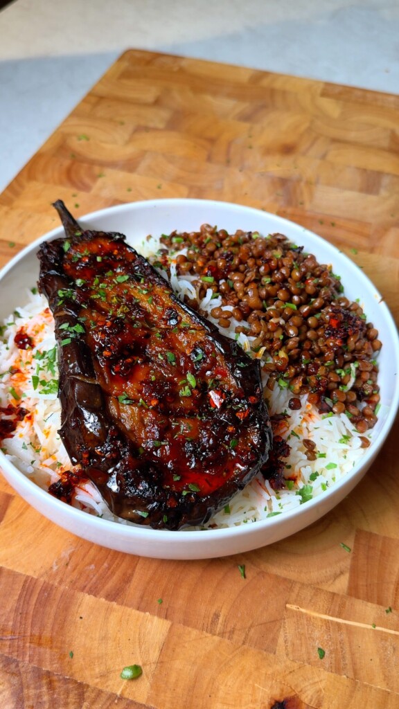 Glazed roasted eggplant served over fluffy white rice with seasoned lentils, garnished with chopped herbs and chilli flakes in a white bowl on a wooden surface.