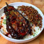Glazed roasted eggplant served over fluffy white rice with seasoned lentils, garnished with chopped herbs and chilli flakes in a white bowl on a wooden surface.