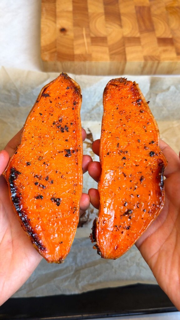 Two roasted sweet potato halves with caramelized cut sides held over a parchment-lined tray