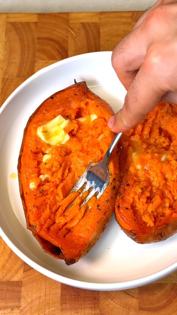 Fork mashing roasted sweet potato flesh with melting vegan butter in a white bowl