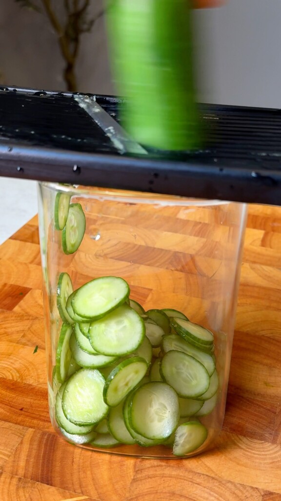 Thinly sliced cucumber falling into a glass jar on a mandoline