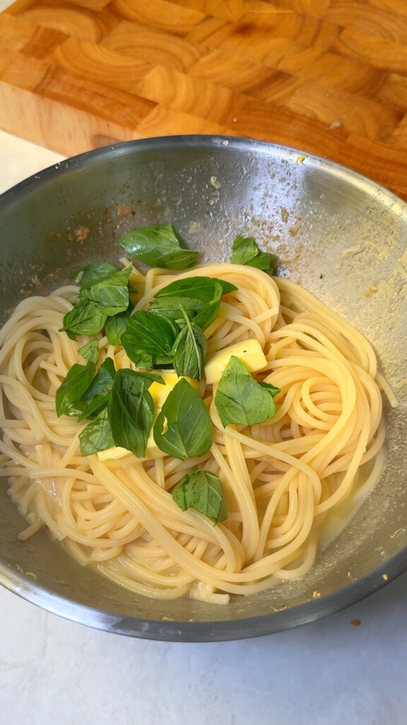 Cooked spaghetti tossed with cubes of vegan butter and fresh basil leaves in a mixing bowl