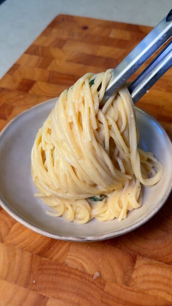 Tongs lifting a twirl of creamy lemon spaghetti from a serving bowl