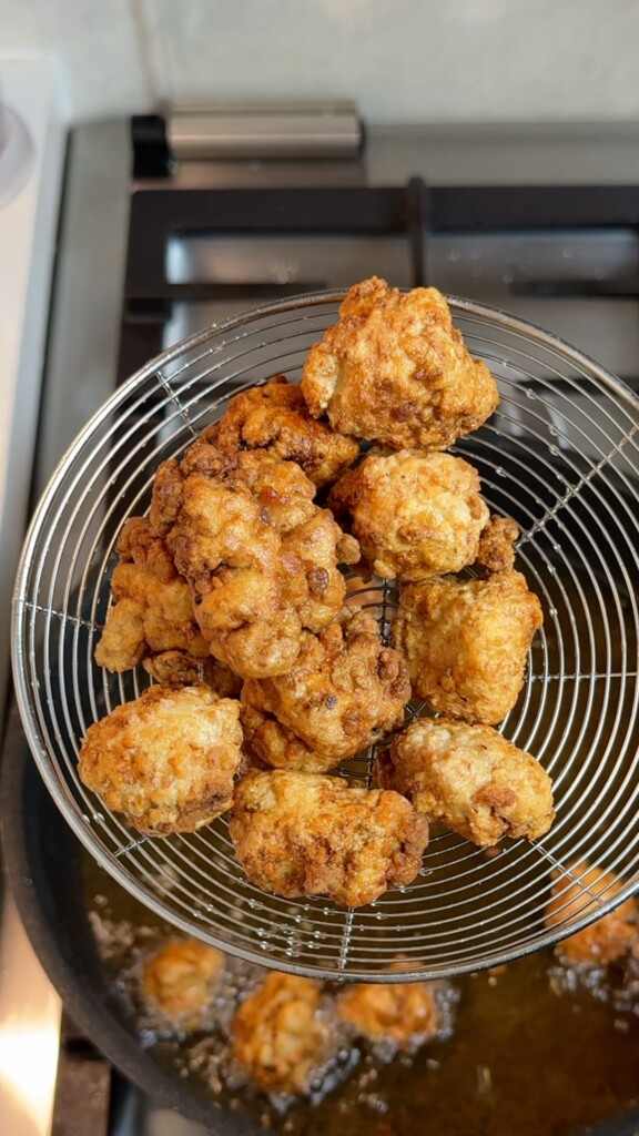 Golden crispy fried popcorn tofu pieces in a spider strainer lifted over a pot of hot oil