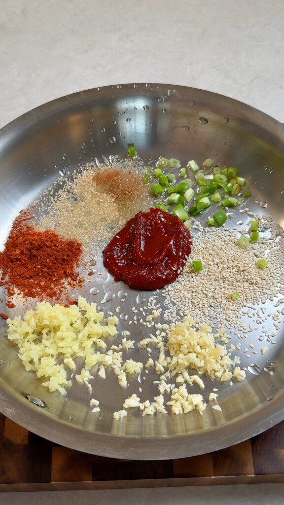 Gochujang paste, gochugaru, sugar, sesame seeds, minced garlic, ginger, and sliced spring onions arranged in a stainless steel pan