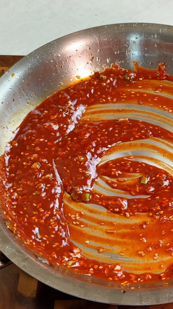 Glossy red gochujang sauce with sesame seeds and spring onions swirled in a stainless steel pan