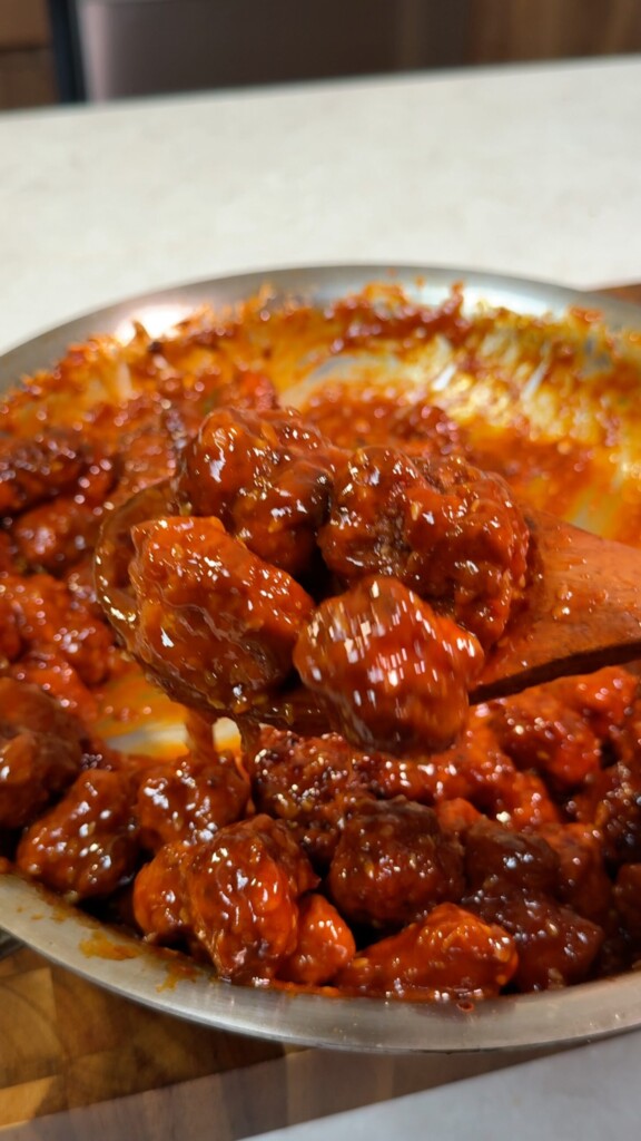 Wooden spoon lifting sticky glazed gochujang tofu coated in spicy red sauce from the pan