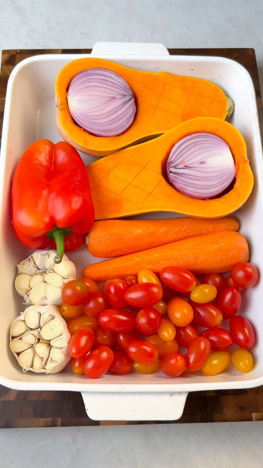 Halved butternut squash with red onion, bell pepper, carrots, garlic heads and cherry tomatoes in a roasting dish