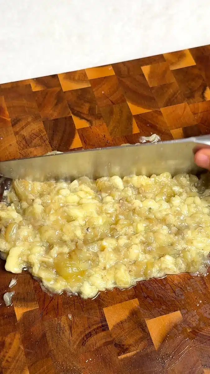 Roasted eggplant flesh being chopped with a knife on a wooden board