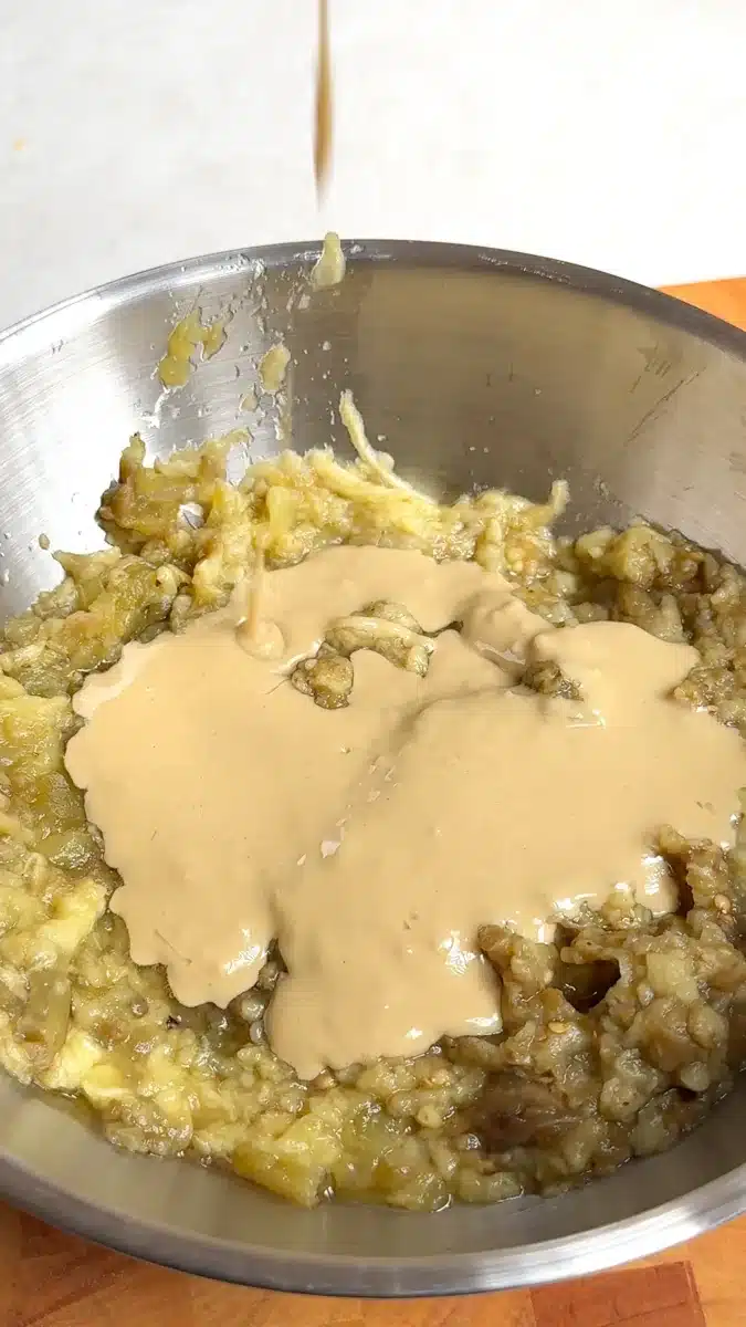 Tahini being poured over chopped roasted eggplant in a mixing bowl