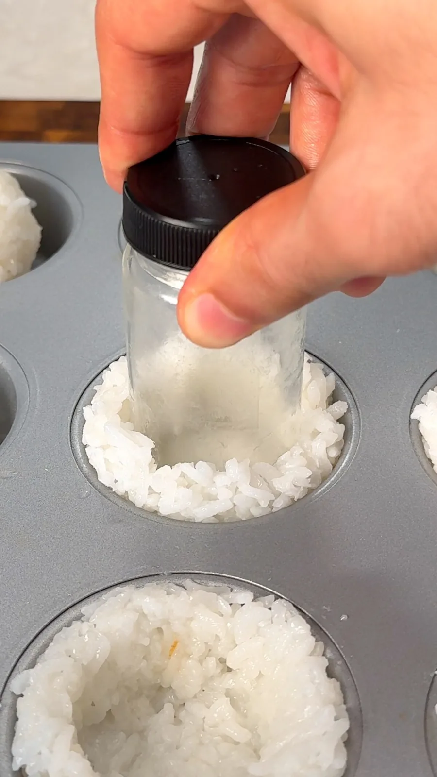 Sushi rice being pressed into a muffin tin with a small jar to form cups