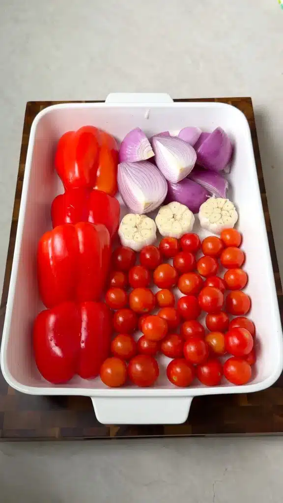 Red pepper halves, onion, garlic heads, and cherry tomatoes arranged in a roasting dish