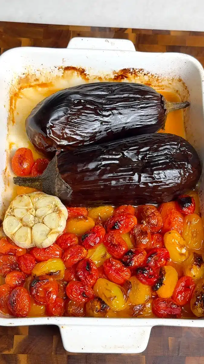 Whole roasted eggplants, cherry tomatoes and a cut garlic bulb in a baking dish