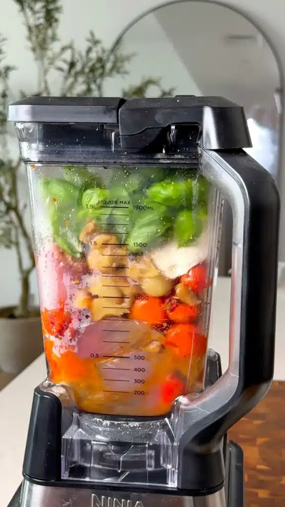 Blender filled with roasted vegetables, silken tofu, garlic, and fresh basil