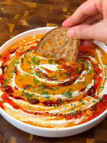 Sourdough bread dipped into a bowl of vegan roasted butternut squash soup with coconut milk and chili oil swirl
