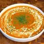 Roasted tomato and red pepper soup in a white bowl with a swirl of vegan cream and chopped herbs