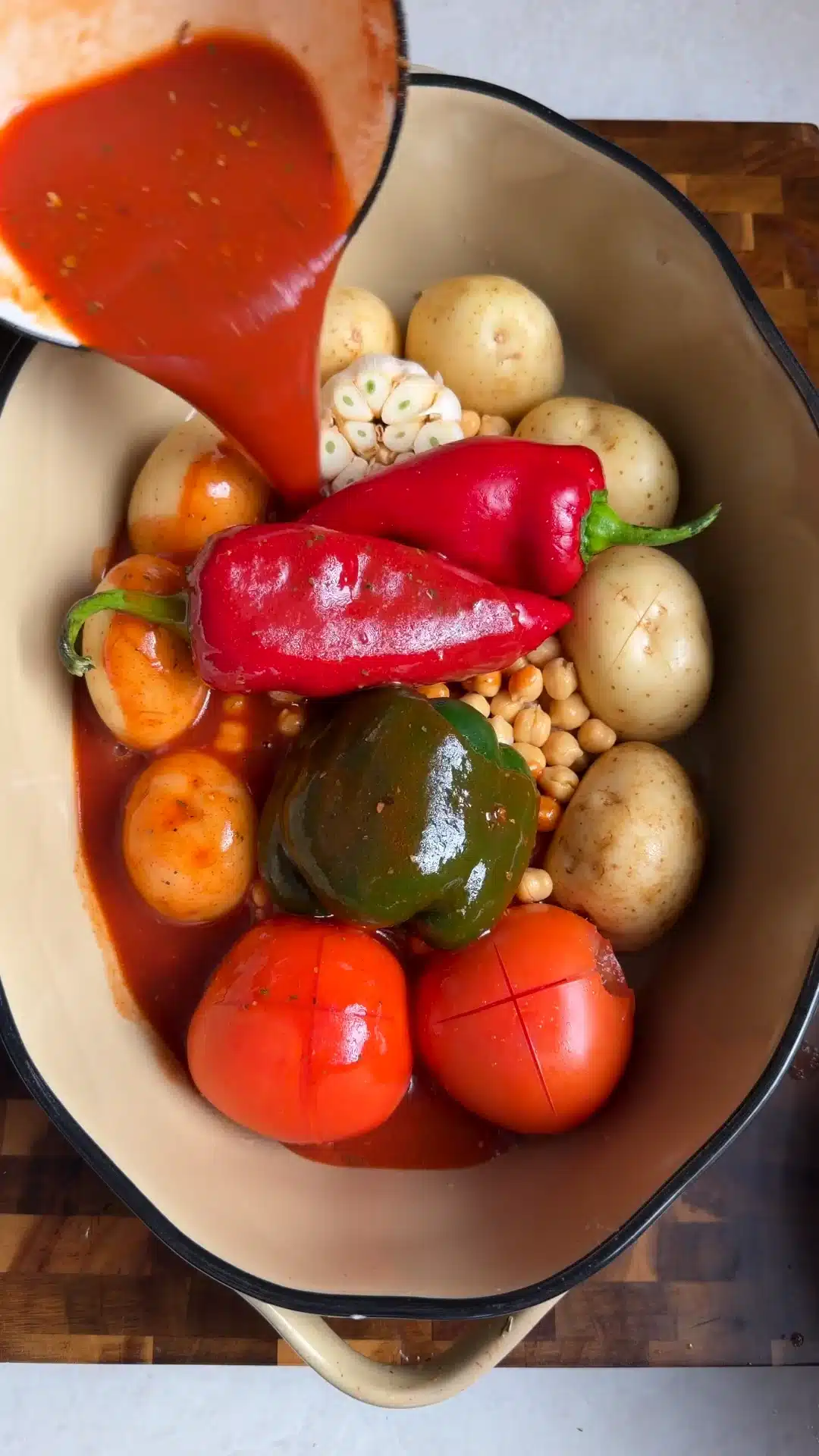 Tomato sauce being poured over small potatoes, chickpeas, peppers, tomatoes and a garlic bulb in a roasting dish.